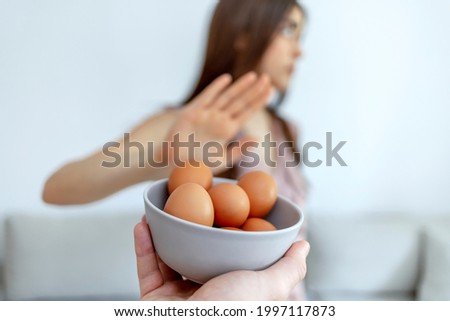Selective focus of a beautiful young women  refusing to eat eggs.  Young beautiful woman holding fresh egg at home with open hand doing stop sign with serious and confident expression. 