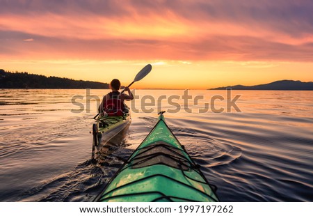 Woman on a sea kayak is paddling in the ocean. Colorful Sunset Sky Art Render. Taken in Jericho, Vancouver, British Columbia, Canada. Royalty-Free Stock Photo #1997197262