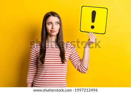 Portrait of attractive doubtful girl holding exclamation point card deciding isolated over bright yellow color background