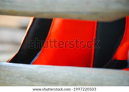 Closeup of red life jacket with black lining inside is hanging behind white wooden balcony. Sea travel safety concept. Empty space at the vest chest can use as mockup.