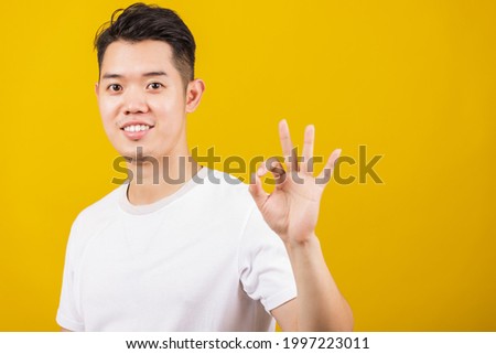 Asian handsome young man smiling positive holding ok sign gesturing with hand and fingers, studio shot isolated on yellow background, making successful expression gesture concept