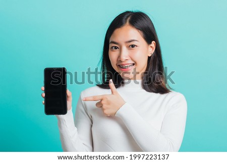 Beautiful Asian woman smile holding a smartphone on hand and pointing finger to the blank screen, female excited cheerful her show mobile phone isolated on a blue background, Technology concept
