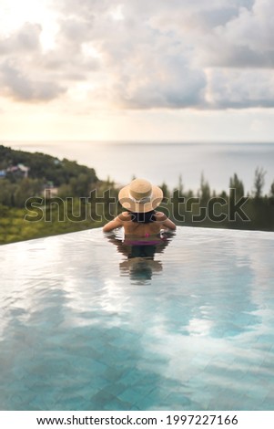 Summer travel on holiday at southeast tropical concept. Rear view asia woman relax at outdoor pool resort sea panorama view. With straw hat and dramatic sky. Phuket island, Thailand