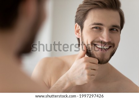 Happy millennial young handsome man looking in mirror in bathroom with toothy smile, scanning facial skin and stylish stubble. Guy satisfied with male beauty care product. Skincare, home spa concept Royalty-Free Stock Photo #1997297426