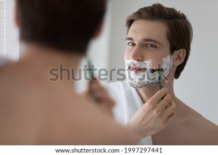 Positive attractive young guy with gel foam on lower face shaving with razor at mirror. Handsome man enjoying morning bath routine, caring for appearance, healthy facial skin. Hygiene, skincare Royalty-Free Stock Photo #1997297441