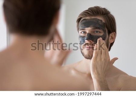 Focused handsome metrosexual guy applying dark cleansing natural clay or mud cosmetic mask on face at mirror for skin treatment, cleaning pores, preventing wrinkles, good complexion. Skincare concept Royalty-Free Stock Photo #1997297444