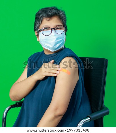 Asian middle age woman wearing protective hygiene mask showing bandage on arm looking to camera with happy trust and confidece after getting vaccine for protect coronavirus or covid-19.