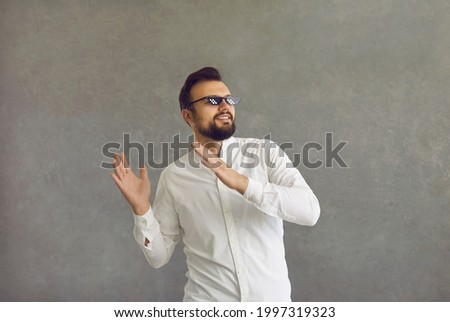 Happy young man in thug life glasses dancing isolated on a grey background. Portrait of a cheerful positive guy in funny sunglasses having fun and dancing to music in the studio