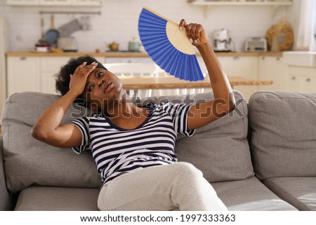 Tired African American woman suffering from heatstroke flat without air-conditioner, waving blue fan, sitting on sofa at home. Black girl cooling in hot summer weather. Overheating, high temperature. Royalty-Free Stock Photo #1997333363