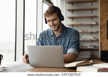 Smiling young handsome man in wireless headphones listening educational webinar lecture on computer, writing notes, improving professional knowledge, studying on online courses in modern home office. Royalty-Free Stock Photo #1997387924