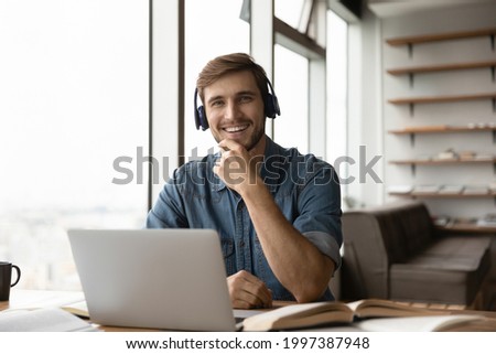 Portrait of happy millennial generation man in headphones sitting at table with books and computer. Smiling young male student posing in modern home office, e-learning distantly on online courses. Royalty-Free Stock Photo #1997387948