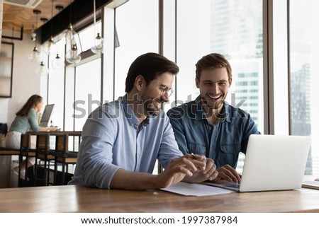 Happy sincere friendly young male colleagues laughing, holding pleasant conversation or watching funny video, enjoying break pause timer working on online project on computer in modern office room. Royalty-Free Stock Photo #1997387984