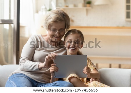 Happy loving middle aged granny resting on comfortable sofa with joyful adorable preschool kid girl, using digital computer tablet together, watching funny photo video content or playing online games.