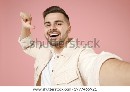 Close up young confident excited fun caucasian man 20s in jacket white t-shirt doing selfie shot on mobile phone point thumb finger on himself isolated on pastel pink color background studio portrait