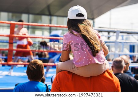 A little girl sits on her father's neck and watches a boxing competition. Street fights in the ring. Unrecognizable people