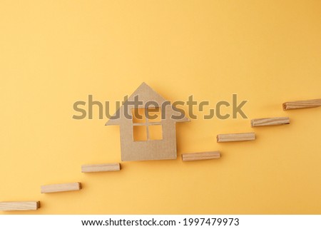 View from above of a house model made from card board cut out on the yellow background with wooden sticks forming stairs