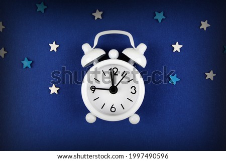A white alarm clock on a blue background, decorated with white and blue stars around.The concept of sleep time, Christmas composition,insomnia, reminder, starfall, romantic date.Flatlay.Copyspace