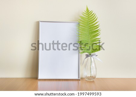 Mockup template of vertical A4 frame standing next to wall with green forest leaf in glass vase.