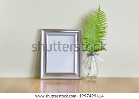 Blank metallic silver frame with blank space and wild forest fern leaf in glass vase on wooden surface.