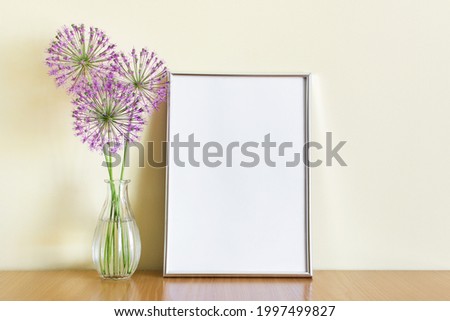 Mockup template with A4 frame standing on wooden shelf with three purple summer flowers branches in glass vase.