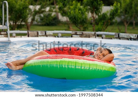 A cute brunette girl 8 years old relaxes by swimming in a frame pool on a swimming circle in the summer in the backyard of a private house.