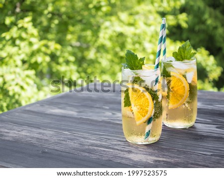 Cold carbonated lemonade with ice, lemon, currant leaves and a cocktail tube in a transparent glass. Summer drink stands on a wooden table in the garden. Background - green tree branches