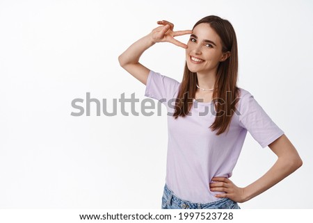 Online positive vibes. Happy beautiful woman 25s years, showing peace disco v-sign near eye, smiling and looking lovely at camera, standing against white background
