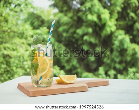 Cold lemonade with ice and lemon on a wooden board. Summer drink in the garden. The background is green tree branches.