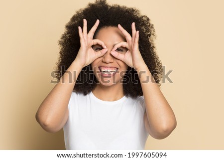 Headshot portrait of excited young African American woman isolated on yellow studio background have fun make glasses with fingers. Profile picture of smiling biracial female with funny facial gesture.