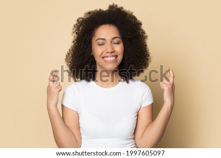 Smiling young African American woman isolated on yellow studio background cross fingers make wish. Happy millennial mixed race female feel excited optimistic for dream come true. Luck concept.