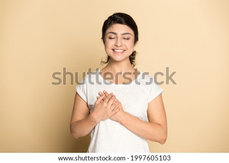Happy religious millennial Indian female isolated on yellow studio background hold hands at heart chest feeling grateful. Thankful young mixed race woman show love gratitude. Faith, believe concept.