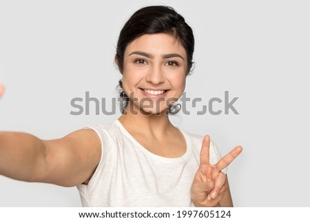 Smiling Indian young woman isolated on grey studio background look at camera make self-portrait picture. Happy millennial mixed race female pose with victory hand gesture take selfie on modern gadget.