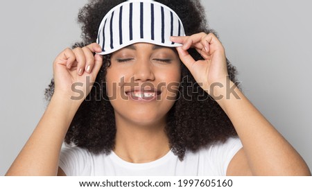 Close up of happy young African American woman isolated on grey studio background wear eye mask go to sleep. Smiling millennial mixed race female get ready for nap or daydream. Relaxation concept.