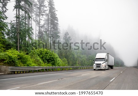 White big rig long haul semi truck transporting commercial cargo in dry van semi trailer running on turning one way highway road with forest on the hill at strong fog weather with poor visibility  Royalty-Free Stock Photo #1997655470