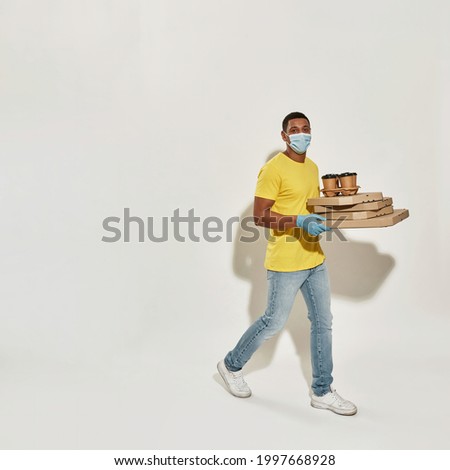 Full length shot of delivery man wearing protective mask and gloves looking at camera, holding many pizza boxes and drinks isolated over gray background. Food delivery service
