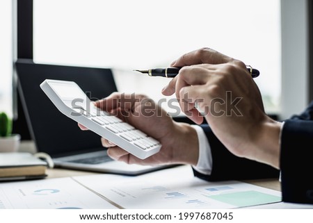 A man with a pen pointing to a white calculator and documents are placed on the table, a businessman in a black suit is sitting to check information from documents, he is a company executive.