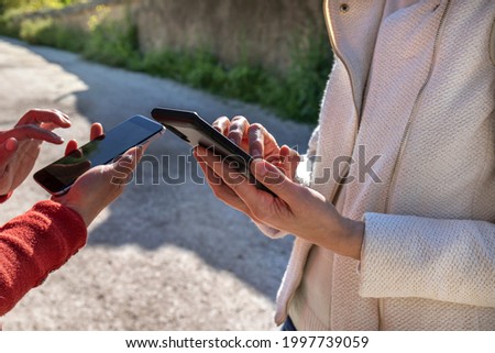 Close up of two girls' hands using their smartphone in a park. One girl wears a white jacket and the other one a red sweater. A country lane with a wall on the background. 