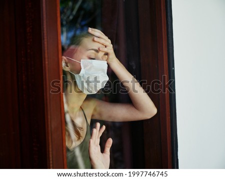 woman with closed eyes in medical mask holds hand on forehead near lockdown window