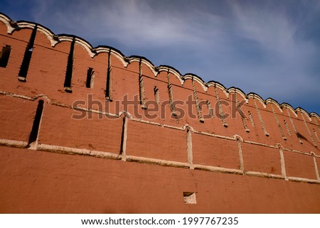 Upper part of a red stone wall in a medieval Russian monastery with blue sky on background.