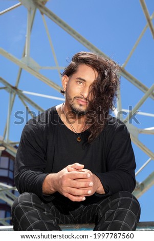 Portrait of young man latin acrobatic street dancer with long hair sitting in a skate park.