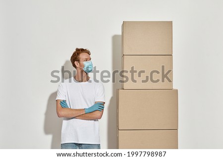 Picture of a young courier with hands folded watches on the tower of cardboard boxes, deliveryman assess the scope of work, wears medical mask and gloves