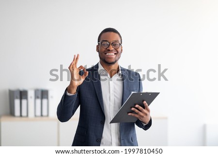 Portrait of cheerful black male psychologist showing okay gesture, recommending professional help at clinic. Confident young African American psychotherapist smiling at camera in office
