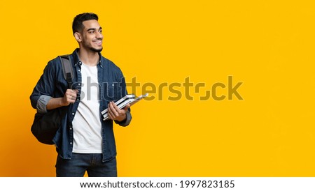 Positive arab guy student with backpack and bunch of books looking at copy space for advertisement over yellow studio background, panorama. Education, university, college, studying, course concept