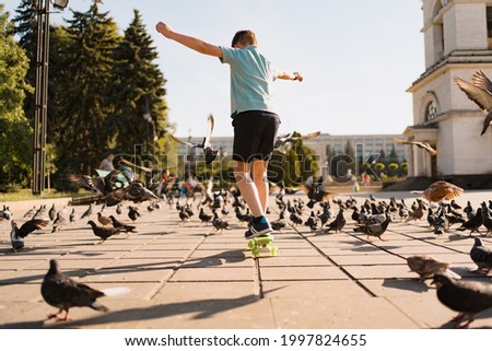 A boy riding penny board in the park on a warm summer time with pigeons and sky