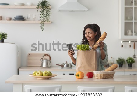 Online buying food and grocery. Delivery service and mobile app. Happy young african american woman sort vegetables from eco package in minimalist kitchen interior, looks at phone, typing message Royalty-Free Stock Photo #1997833382