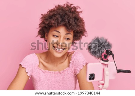 Blogging podcast recording and video streaming concept. Smiling Afro American woman has natural curly hair looks at smartphone camera talks with followers isolated over pink studio background.