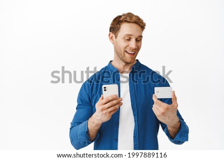 Attractive man with red hair, paying with mobile phone and credit card, looking happy while entering banking number into smartphone app, make delivery order, standing over white background