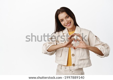 Beautiful tender girl shows heart sign, woman make I love you gesture and smiling cute, express her feelings, standing in casual clothes against white background