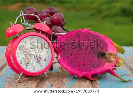 Red grapes , dragon fruit  and alarm clock on rustic wooden with green field background , fresh fruit concept