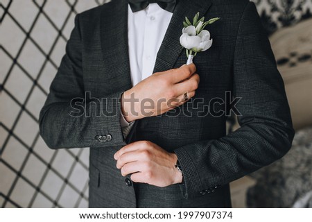 A man, a groom in a gray checkered suit straightens his boutonniere and bow-tie with his hand. Wedding portrait.